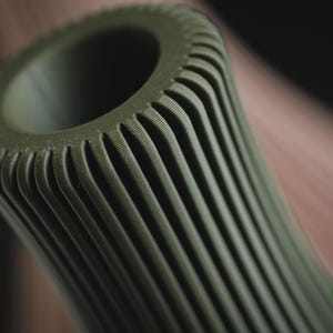 May include: An olive green cylindrical object with a hollow centre and a series of vertical, parallel ridges running along its exterior. The surface features a fine, textured finish. A small circular logo with the text h3li&oslash; design is visible.