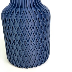 May include: A matte navy blue vase featuring a prominent geometric diamond lattice pattern across its cylindrical body. The structure transitions into vertical ridges near the neck, creating a textured surface with consistent depth and symmetry.