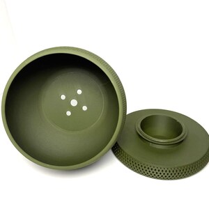 Puede incluir: Una maceta redonda de pl&aacute;stico de color verde oliva, compuesta por dos piezas con un exterior texturizado de patr&oacute;n de diamantes. El cuenco principal tiene cinco orificios de drenaje en la base y la base circular separada tiene un anillo central.