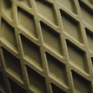 May include: A close-up view of a matte olive green object featuring a repeating diamond-shaped lattice pattern with visible 3D printed layer lines. A small circular logo in the bottom right corner displays the text h3li&oslash; design.