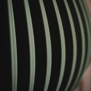 May include: A spherical object featuring a series of vertical, ribbed panels in a muted sage green colour, separated by deep black gaps. The surface texture appears finely lined and structured. A small circular logo with the text h3li&oslash; design is visible.