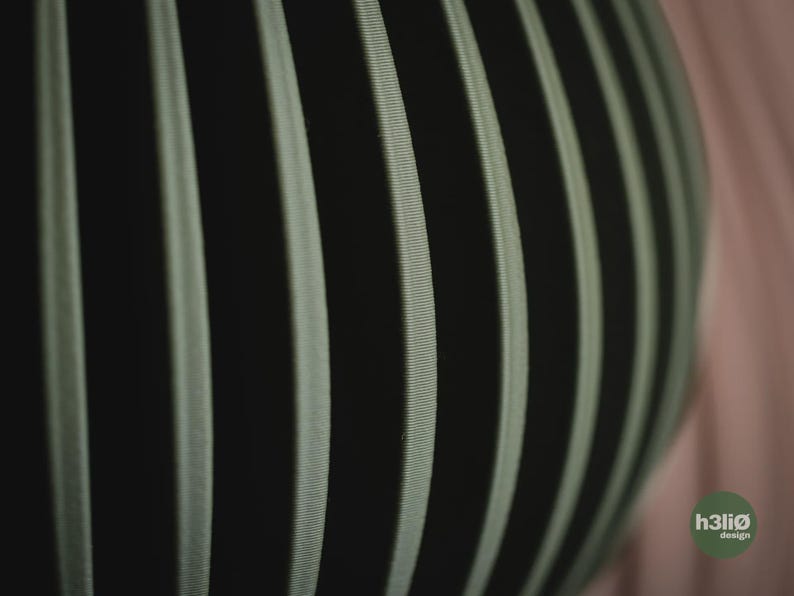 May include: A spherical object featuring a series of vertical, ribbed panels in a muted sage green colour, separated by deep black gaps. The surface texture appears finely lined and structured. A small circular logo with the text h3li&oslash; design is visible.