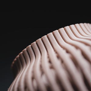 K&ouml;nnte beinhalten: Ein hellbeiges, dreidimensionales Objekt mit einer sich wiederholenden, wellenf&ouml;rmigen, vertikal gerippten Textur. Die Oberfl&auml;che weist feine, horizontale Schichtlinien auf, die f&uuml;r die additive Fertigung typisch sind. Ein kleines gr&uuml;nes kreisf&ouml;rmiges Logo zeigt h3li&oslash; design.