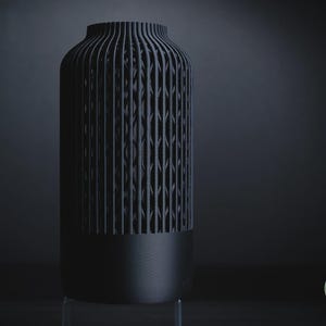 K&ouml;nnte beinhalten: Eine mattschwarze zylindrische Vase mit einem massiven Boden und einem oberen Bereich mit vertikalen Lamellen und einem geometrischen Gittermuster. Der Text h3li&oslash; design ist in einem kleinen gr&uuml;nen Kreis unten rechts zu sehen.