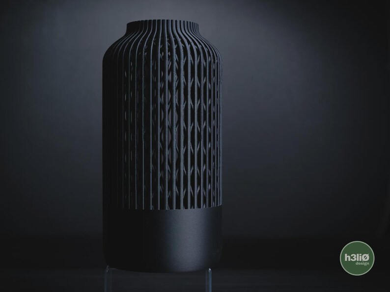 K&ouml;nnte beinhalten: Eine mattschwarze zylindrische Vase mit einem massiven Boden und einem oberen Bereich mit vertikalen Lamellen und einem geometrischen Gittermuster. Der Text h3li&oslash; design ist in einem kleinen gr&uuml;nen Kreis unten rechts zu sehen.