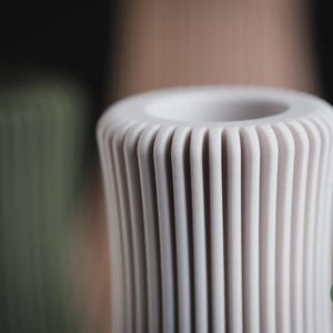May include: A cylindrical off-white vase featuring a series of vertical, rounded ridges along its exterior surface. The top of the vase has a circular opening. A small green circular logo with the text h3li&oslash; design is visible in the corner.