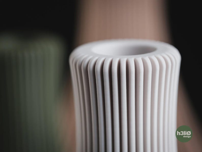 May include: A cylindrical off-white vase featuring a series of vertical, rounded ridges along its exterior surface. The top of the vase has a circular opening. A small green circular logo with the text h3li&oslash; design is visible in the corner.