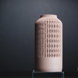 K&ouml;nnte beinhalten: Eine zylindrische Vase in sanftem Pfirsichton mit einem vertikalen Gittermuster auf den oberen zwei Dritteln des K&ouml;rpers. Der Boden ist massiv und glatt, w&auml;hrend der obere Rand gerippt ist. Ein kleines kreisf&ouml;rmiges Logo mit dem Text h3li&oslash; design ist sichtbar.