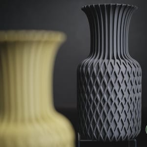May include: A dark grey vase with a vertical ribbed neck and a diamond-patterned body, featuring a circular green logo with the text h3li&oslash; design in the bottom right corner, positioned next to a blurred yellow vase of the same design.