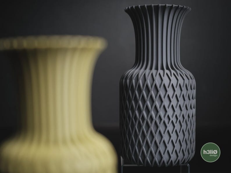 May include: A dark grey vase with a vertical ribbed neck and a diamond-patterned body, featuring a circular green logo with the text h3li&oslash; design in the bottom right corner, positioned next to a blurred yellow vase of the same design.