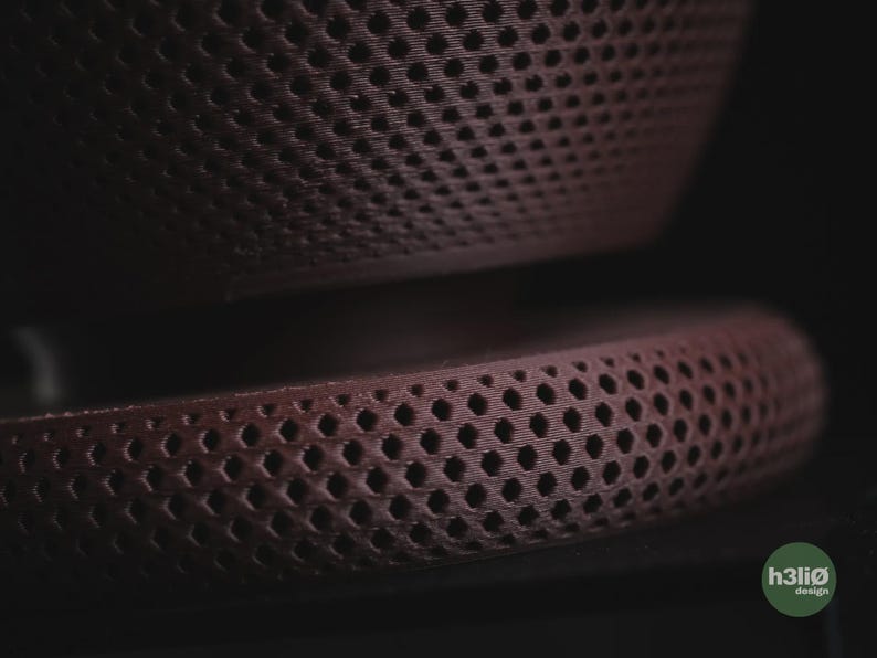 Puede incluir: Un objeto de color granate oscuro con una estructura de celos&iacute;a de panal. La superficie presenta un patr&oacute;n de rejilla hexagonal repetitivo en toda su forma curva. Un peque&ntilde;o logotipo circular verde con el texto h3li&oslash; design es visible.