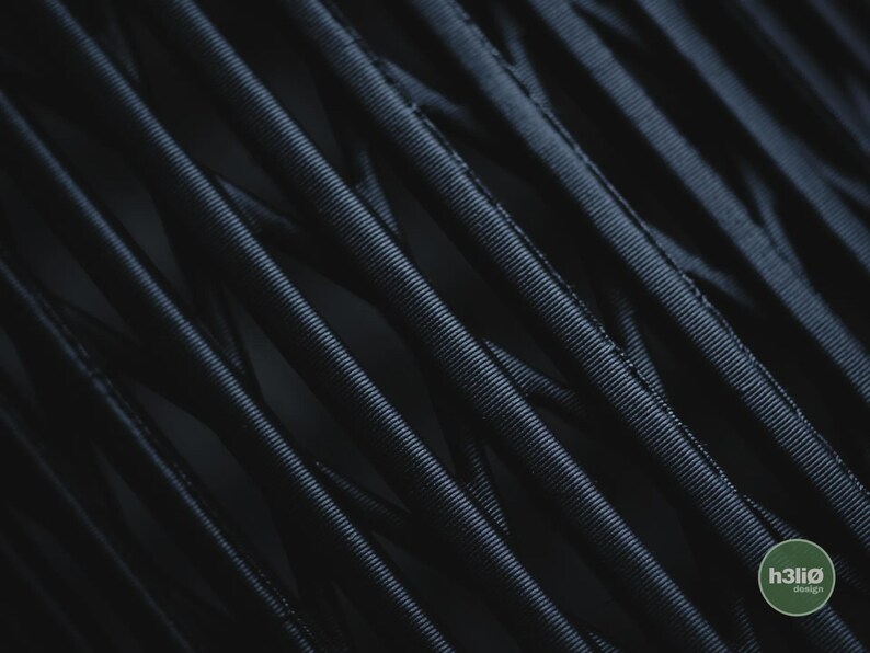 K&ouml;nnte beinhalten: Eine dunkel, mattschwarze 3D-gedruckte Struktur mit einem dichten, sich kreuzenden Gitter aus d&uuml;nnen, gerippten zylindrischen Filamenten. Die Oberfl&auml;chenstruktur ist durch feine, horizontale Rillen entlang jedes Strangs gekennzeichnet. Ein kleines gr&uuml;nes Logo zeigt h3li&oslash; design.