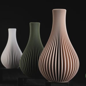 May include: Three vases with a vertical ribbed texture and a tapered neck. The vases are white, olive green, and light peach in colour. Each vase stands on a small black rectangular base. The text h3li&oslash; design is visible in the bottom right corner.