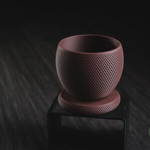 Puede incluir: Una maceta de color malva oscuro mate con cuerpo esf&eacute;rico y base circular a juego, ambos con un patr&oacute;n de celos&iacute;a en forma de diamante en toda la superficie exterior. El texto h3li&oslash; design es visible en la esquina inferior derecha.