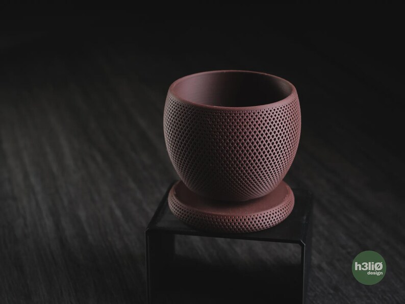Puede incluir: Una maceta de color malva oscuro mate con cuerpo esf&eacute;rico y base circular a juego, ambos con un patr&oacute;n de celos&iacute;a en forma de diamante en toda la superficie exterior. El texto h3li&oslash; design es visible en la esquina inferior derecha.