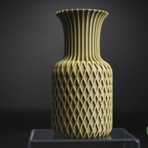 May include: A pale olive green vase with a cylindrical body featuring a raised diamond lattice texture and a flared, vertically ribbed neck. The base of the vase is smooth, and a small circular logo with the text h3li&oslash; design is visible.