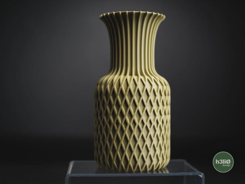 May include: A pale olive green vase with a cylindrical body featuring a raised diamond lattice texture and a flared, vertically ribbed neck. The base of the vase is smooth, and a small circular logo with the text h3li&oslash; design is visible.