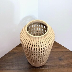 K&ouml;nnte beinhalten: Eine hellbeige Vase in zylindrischer Form mit einer hohlen Gitterstruktur. Die Au&szlig;enseite besteht aus vertikalen, geschwungenen Rippen, die sich kreuzen und ein sich wiederholendes Rautenmuster &uuml;ber den gesamten K&ouml;rper des Objekts bilden.