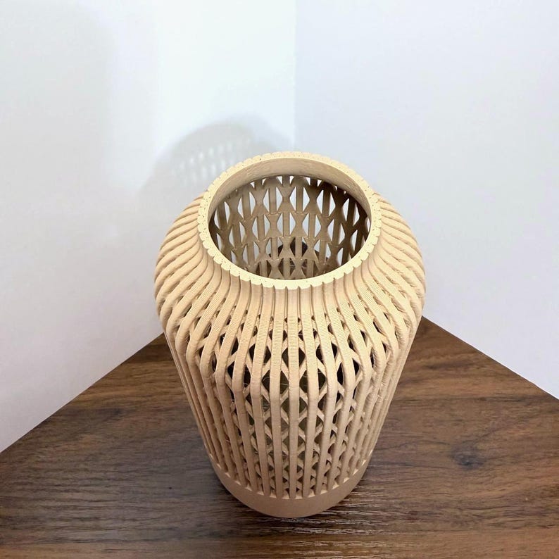 K&ouml;nnte beinhalten: Eine hellbeige Vase in zylindrischer Form mit einer hohlen Gitterstruktur. Die Au&szlig;enseite besteht aus vertikalen, geschwungenen Rippen, die sich kreuzen und ein sich wiederholendes Rautenmuster &uuml;ber den gesamten K&ouml;rper des Objekts bilden.