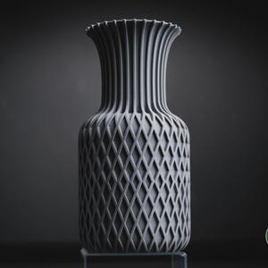 May include: A matte grey vase featuring a vertical ribbed neck and a textured diamond lattice pattern on the body. The base of the vase is cylindrical, and a small circular logo with the text h3li&oslash; design is visible in the bottom right corner.