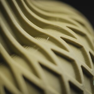 May include: A light olive green object featuring a repeating geometric pattern of raised, wavy ridges and interconnected diamond shapes. The surface texture shows fine, parallel lines from a 3D printing process. A small circular logo reads h3li&oslash; design.
