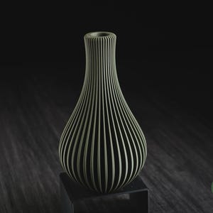 May include: A sage green vase with a vertical ribbed texture and a narrow neck. The vase features a rounded body that tapers upwards to a circular opening. A small green circular logo with the text h3li&oslash; design is visible in the bottom right corner.