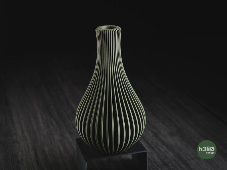 May include: A sage green vase with a vertical ribbed texture and a narrow neck. The vase features a rounded body that tapers upwards to a circular opening. A small green circular logo with the text h3li&oslash; design is visible in the bottom right corner.