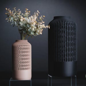 K&ouml;nnte beinhalten: Eine hellrosa Vase und eine schwarze Vase, beide zylindrisch geformt mit einer vertikalen, gitterartigen Struktur im oberen Bereich und einem massiven Sockel. Die hellrosa Vase enth&auml;lt einen Zweig mit gr&uuml;nen Eukalyptusbl&auml;ttern. Der Text h3li&oslash; design ist sichtbar.