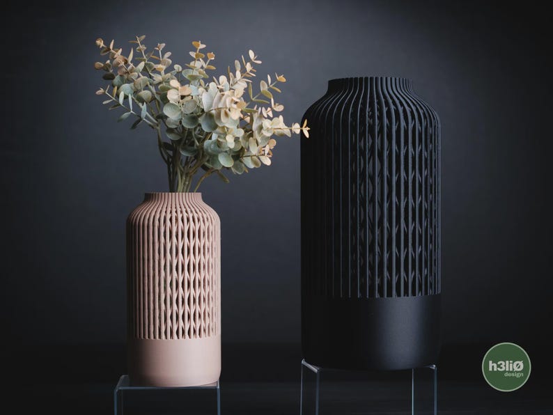 K&ouml;nnte beinhalten: Eine hellrosa Vase und eine schwarze Vase, beide zylindrisch geformt mit einer vertikalen, gitterartigen Struktur im oberen Bereich und einem massiven Sockel. Die hellrosa Vase enth&auml;lt einen Zweig mit gr&uuml;nen Eukalyptusbl&auml;ttern. Der Text h3li&oslash; design ist sichtbar.