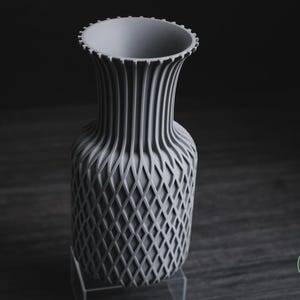 May include: A light grey vase with a textured lattice pattern on the lower body and vertical ribs extending up to the flared, notched rim. The base of the vase features a small circular logo with the text h3li&oslash; design in white lettering.