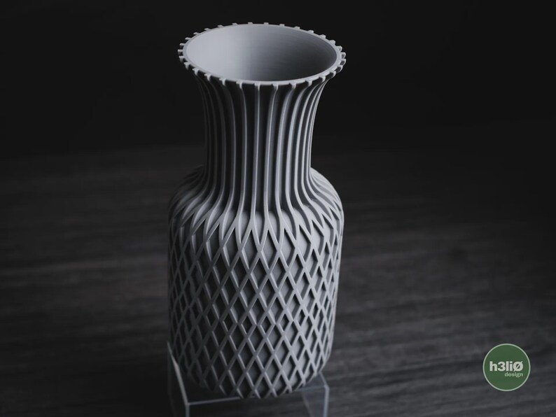 May include: A light grey vase with a textured lattice pattern on the lower body and vertical ribs extending up to the flared, notched rim. The base of the vase features a small circular logo with the text h3li&oslash; design in white lettering.