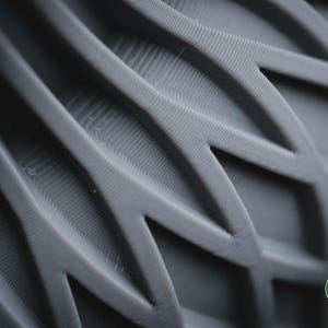 May include: A dark grey 3D printed object featuring a repeating geometric pattern of interconnected, angular, leaf-like shapes with visible horizontal layer lines. A small circular green logo with the text h3li&oslash; design is in the corner.