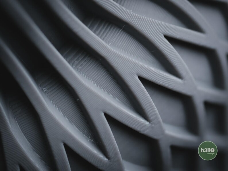 May include: A dark grey 3D printed object featuring a repeating geometric pattern of interconnected, angular, leaf-like shapes with visible horizontal layer lines. A small circular green logo with the text h3li&oslash; design is in the corner.