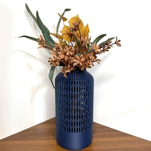 K&ouml;nnte beinhalten: Eine dunkelblaue zylindrische Vase mit einem vertikalen Gittermuster im oberen Bereich und einem massiven Boden. Die Vase enth&auml;lt ein Arrangement aus getrockneten botanischen Elementen, darunter lange gr&uuml;ne Bl&auml;tter und B&uuml;schel kleiner brauner und gelber Bl&uuml;ten.