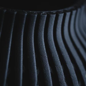 K&ouml;nnte beinhalten: Eine mattschwarze, 3D-gedruckte Vase mit einer Reihe vertikaler, gerippter Stege, die eine texturierte, wellenf&ouml;rmige Oberfl&auml;che bilden. Das Objekt zeigt ein deutliches Schichtmuster und enth&auml;lt ein kleines kreisf&ouml;rmiges Logo mit der Aufschrift h3li&oslash; design.
