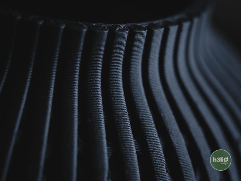 K&ouml;nnte beinhalten: Eine mattschwarze, 3D-gedruckte Vase mit einer Reihe vertikaler, gerippter Stege, die eine texturierte, wellenf&ouml;rmige Oberfl&auml;che bilden. Das Objekt zeigt ein deutliches Schichtmuster und enth&auml;lt ein kleines kreisf&ouml;rmiges Logo mit der Aufschrift h3li&oslash; design.