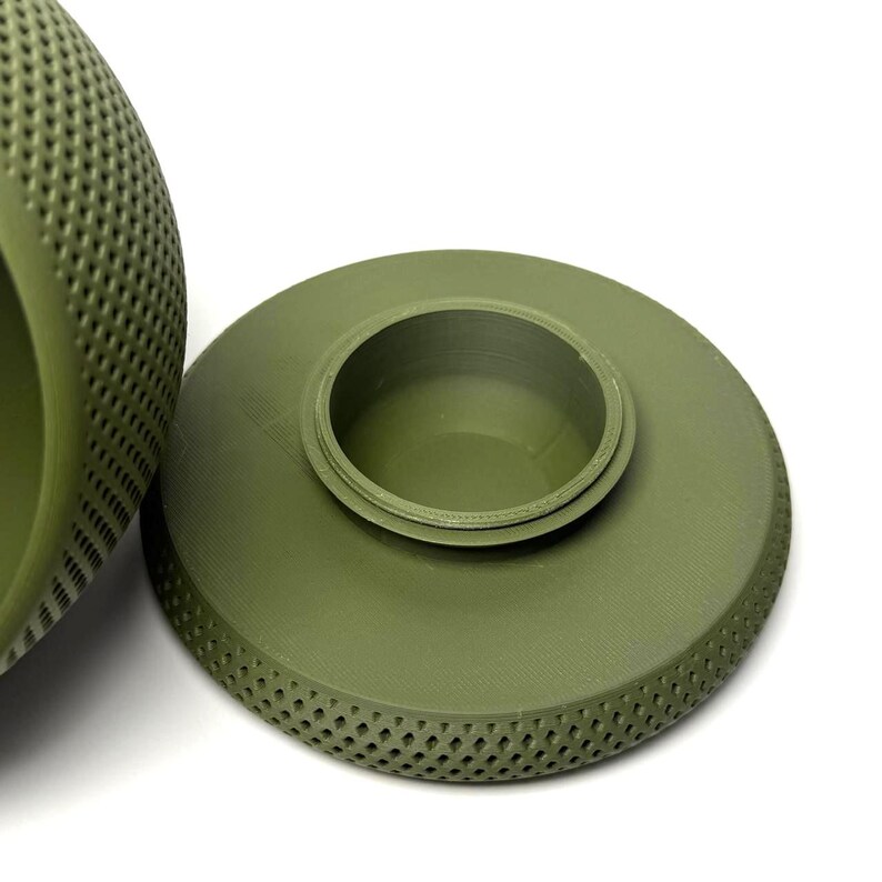 Puede incluir: Un recipiente redondo de color verde oliva con una tapa circular a juego. La tapa presenta un borde interior circular elevado para un cierre seguro, mientras que la superficie exterior tanto del recipiente como de la tapa muestra un patr&oacute;n de rejilla en forma de diamante.