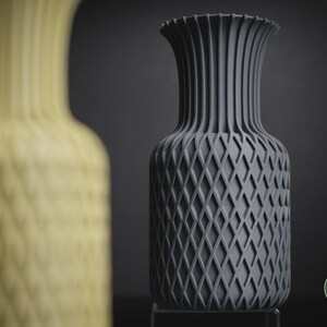 May include: A dark grey vase with a textured diamond lattice pattern on the body and vertical ribs on the neck. The vase features a circular logo with the text h3li&oslash; design in the bottom right corner. A blurred yellow vase is in the foreground.