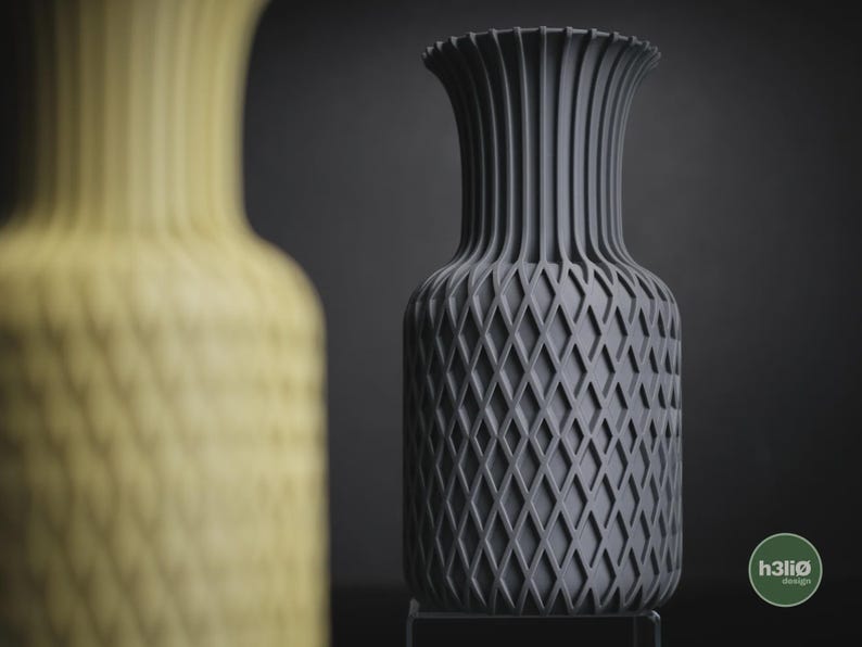 May include: A dark grey vase with a textured diamond lattice pattern on the body and vertical ribs on the neck. The vase features a circular logo with the text h3li&oslash; design in the bottom right corner. A blurred yellow vase is in the foreground.