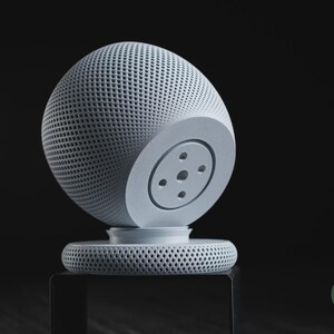 Puede incluir: Un altavoz inteligente esf&eacute;rico de color gris claro con un exterior de malla texturizada y una base circular plana. El dispositivo presenta un panel circular liso y s&oacute;lido en el lateral con cuatro peque&ntilde;as hendiduras. Un logo verde dice h3li&oslash; design.