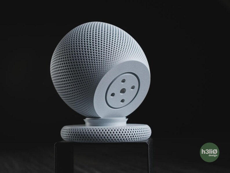 Puede incluir: Un altavoz inteligente esf&eacute;rico de color gris claro con un exterior de malla texturizada y una base circular plana. El dispositivo presenta un panel circular liso y s&oacute;lido en el lateral con cuatro peque&ntilde;as hendiduras. Un logo verde dice h3li&oslash; design.