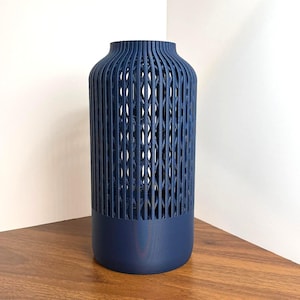 K&ouml;nnte beinhalten: Eine zylindrische marineblaue Vase mit einem massiven Boden und einem oberen Teil mit einer vertikalen, offenen Gitterstruktur. Das Design besteht aus sich wiederholenden d&uuml;nnen, parallelen St&auml;ben, die durch kleine horizontale Segmente verbunden sind.