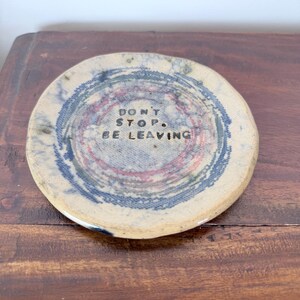 Peut inclure: Un dessous de verre rond en céramique blanc cassé présentant un motif texturé de cercles concentriques dans des tons de bleu et de rouge sourd. Le centre est marqué du texte noir "DON'T STOP. BE LEAVING" en police sans empattement.