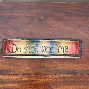 May include: A rectangular ceramic tray with a black rim and a multicoloured interior featuring a rainbow-like gradient of red, yellow, blue, and green. The text "Do not vex me" is written in black, hand-painted lettering along the centre base.