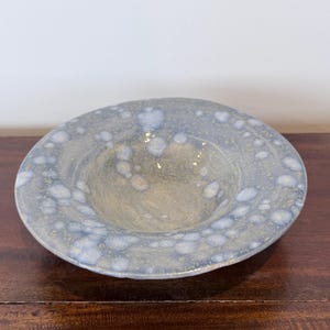 May include: A shallow ceramic bowl with a wide, flat rim featuring a mottled glaze in shades of dusty blue, soft grey, and pale yellow. The interior basin is smooth and concave, while the surface displays a speckled, crystalline texture.