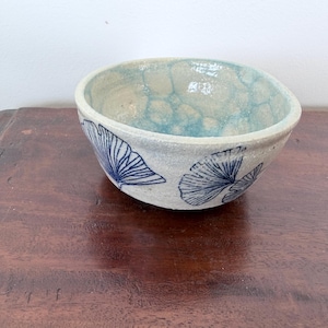May include: A ceramic bowl with a speckled off-white exterior featuring dark blue ginkgo leaf illustrations. The interior of the bowl has a light blue glaze with a textured, bubble-like pattern throughout the surface of the basin.