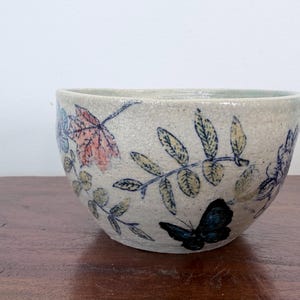 May include: A ceramic bowl with a speckled off-white glaze featuring hand-painted botanical illustrations of leaves in shades of green and yellow, a single red maple leaf, and a prominent black butterfly on the exterior surface.