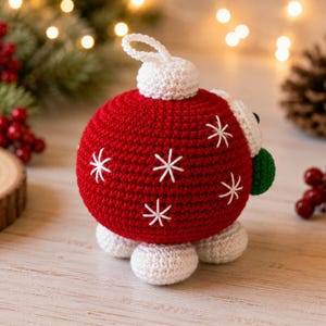 May include: A round, red crocheted ornament featuring white embroidered snowflake patterns. The ornament has a white crocheted top loop, a white base, and three white crocheted feet at the bottom. A small green crocheted accent is visible on the side.