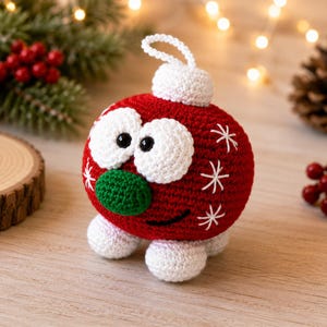 May include: A round, red crocheted ornament featuring two large white eyes with black pupils, a green nose, and a black stitched smile. The ornament has a white top loop, white feet, and white embroidered star-like accents on its sides.