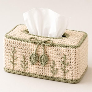 May include: A rectangular crochet tissue box cover in a light beige yarn with a sage green border. The front features embroidered sage green sprigs and a decorative crochet bow with two dangling leaf-shaped accents. A white tissue is visible.